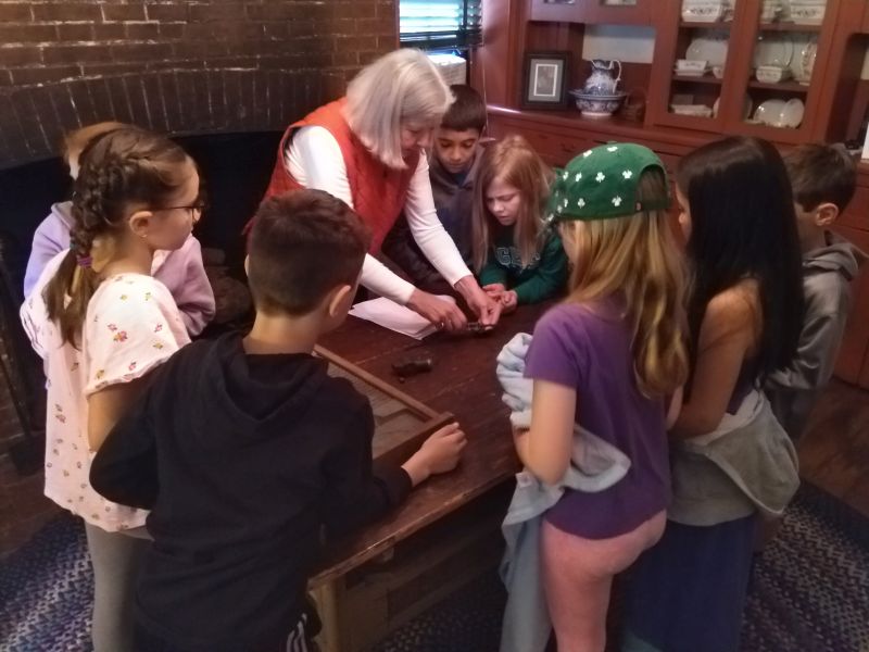 A woman shows a small artifact on a table to a group of students.