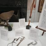 Part of the display from the "Objects That Measure" exhibit. On a table with a white cloth are a trustee postal scale, three calipers of different design, a skirt hem marker, and a micrometer.